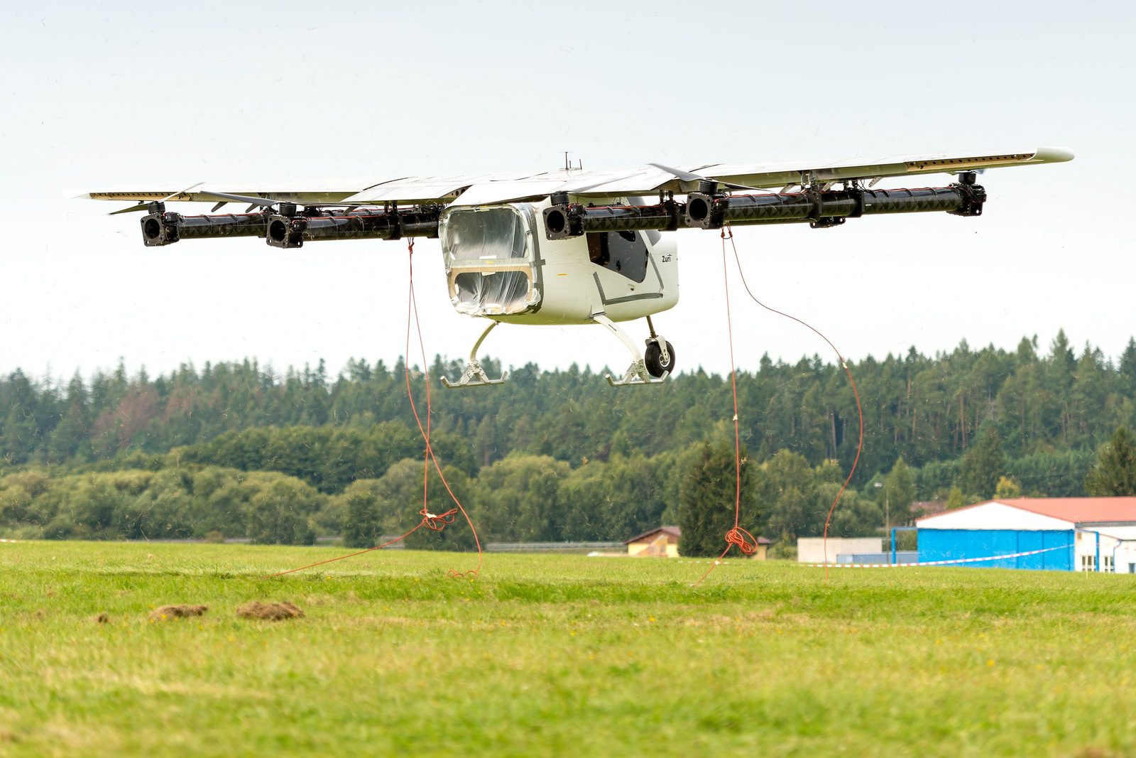 The Zuri TD 1.0 eleven-meter demonstrator airborne on its safety tethers during its first full-scale hover, September 2021, grass airfield and forest behind