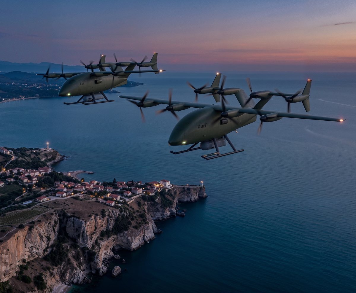 Two Zuri uncrewed cargo aircraft in olive livery flying in loose formation along a coastline after sunset, navigation lights on, a headland town and hills below, a render