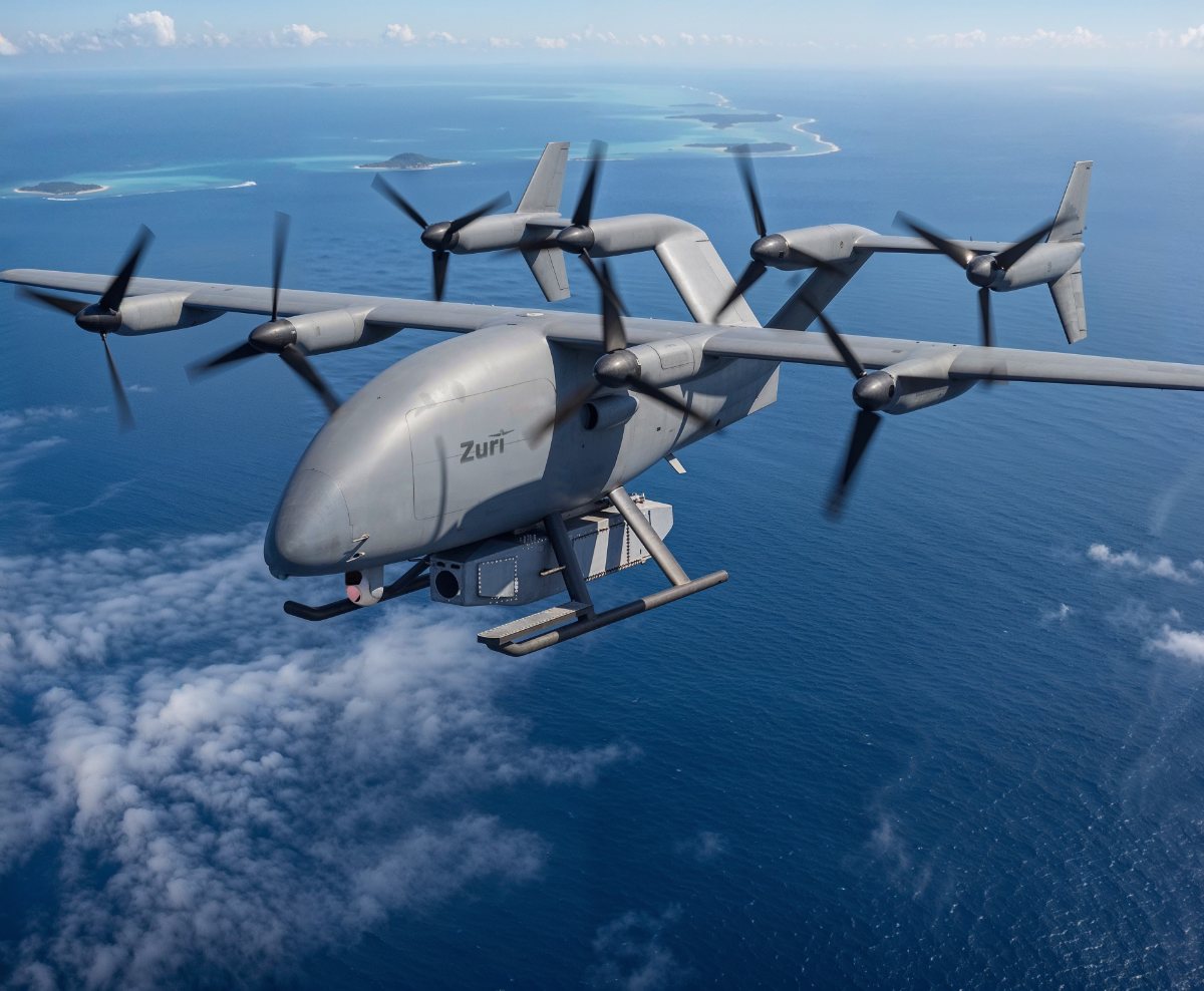 The Zuri uncrewed cargo aircraft in gray livery cruising above open ocean with reef islands beyond, a sensor pod carried under the fuselage, a render