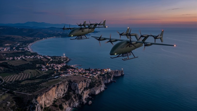 Render of two uncrewed cargo aircraft in formation above a coastline at dusk, a bay and headland below