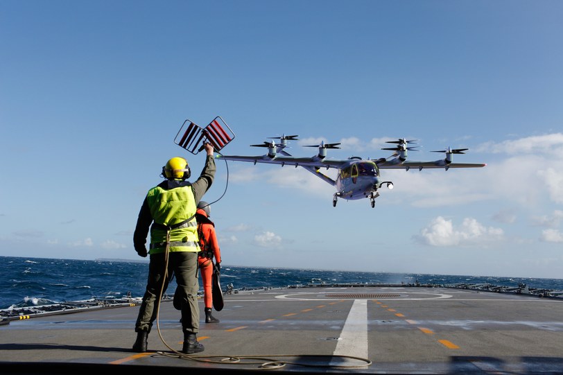 Render of the six-seat aircraft in gray military livery approaching a ship deck, a crew member signaling it in with paddles