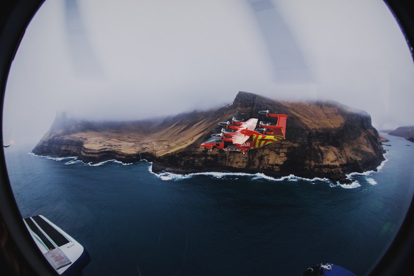 Render looking out through a rounded cabin window at the six-seat aircraft in rescue livery flying low over a rocky island in open sea