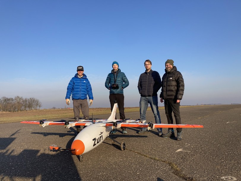 Four members of the Zuri team standing on an airfield behind the V1NG subscale demonstrator, one of them holding a radio control transmitter