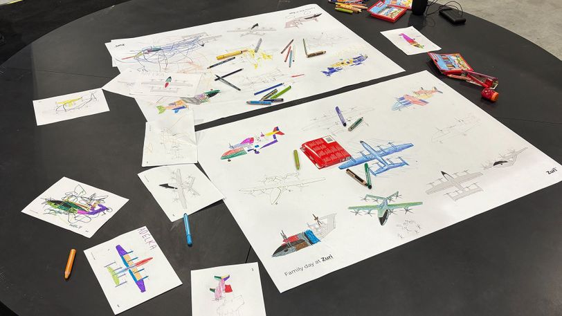 Children's hand-drawn color concepts of the Zuri Hybrid VTOL displayed across the drawing table.