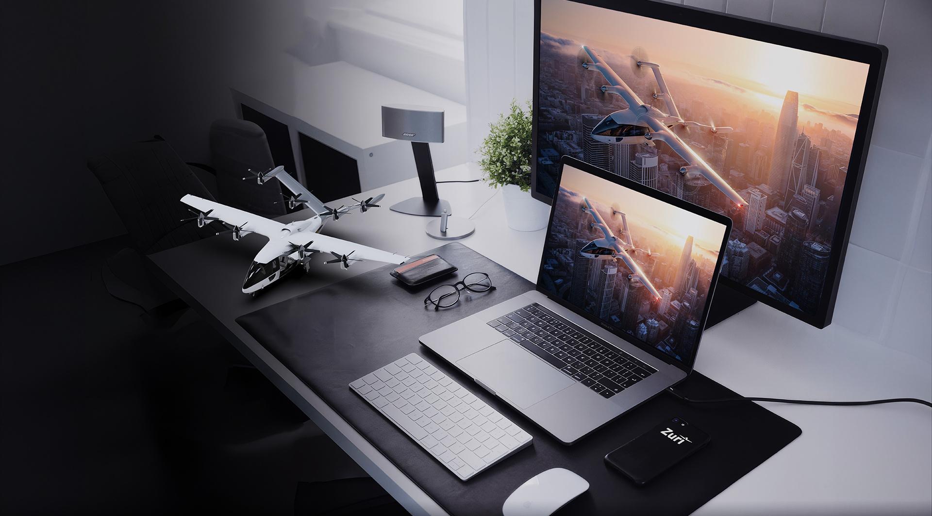 Zuri scale model aircraft displayed on a modern desk setup with a laptop and monitor showing the aircraft in flight.